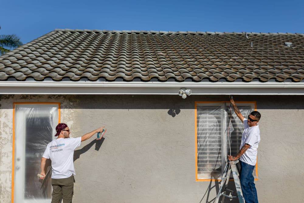 Two workers painting a house exterior with protective coverings on windows and a ladder.