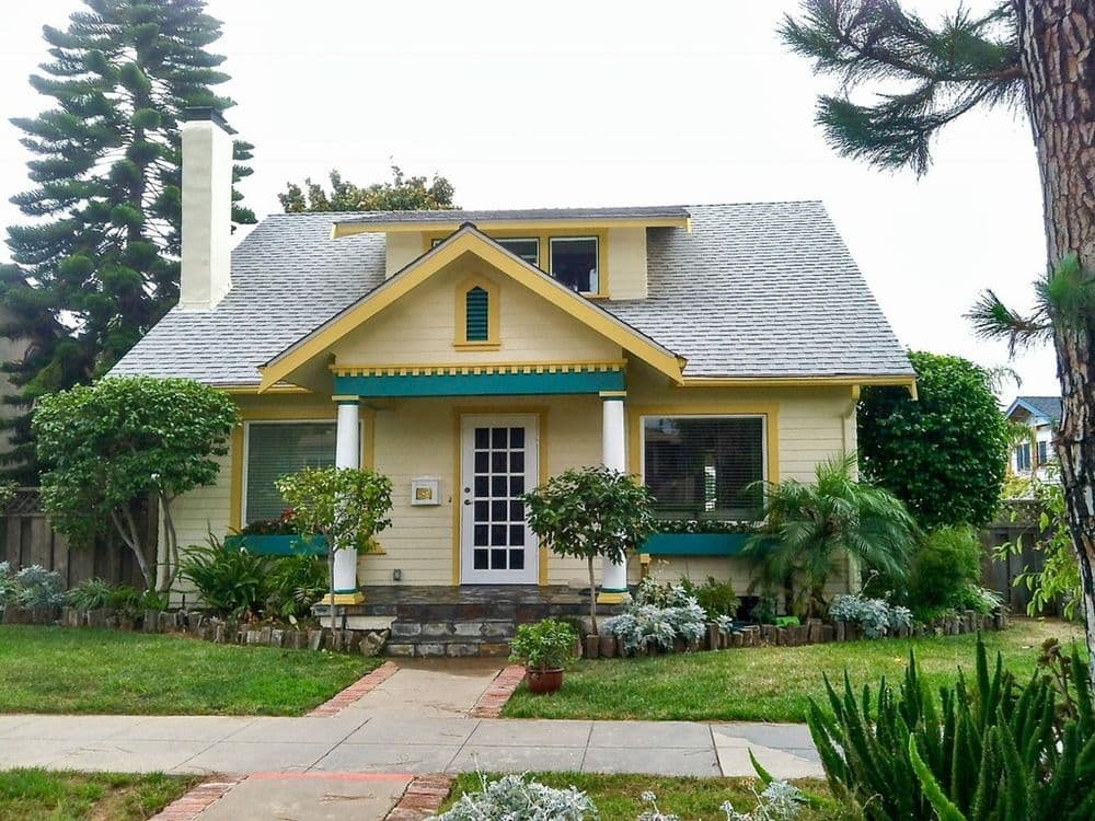Charming yellow house with green accents, surrounded by lush plants and trees.